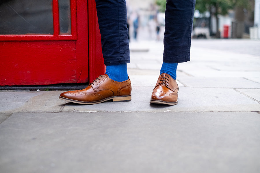 Brown leather shoes and royal blue socks worn on a sidewalk with a red door in the background