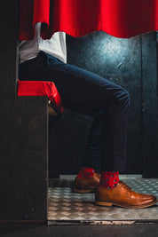 Person wearing brown shoes with red spot socks and dark pants peeking from behind a red curtain.