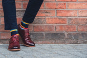Person wearing burgundy shoes and colourful crew socks standing against a brick wall.