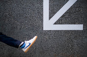 Person wearing white sneakers and bold stripey socks with a blue swoosh logo stepping onto a white arrow on a black asphalt surface.