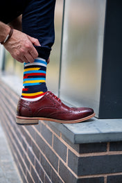 Person wearing a colorful striped premium sock with a brown shoe on a brick wall background