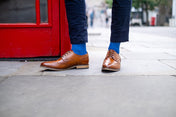 Brown leather shoes and royal blue socks worn on a sidewalk with a red door in the background
