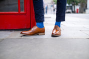 Brown leather shoes and royal blue socks worn on a sidewalk with a red door in the background
