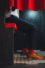 Person wearing brown shoes with red spot socks and dark pants peeking from behind a red curtain.