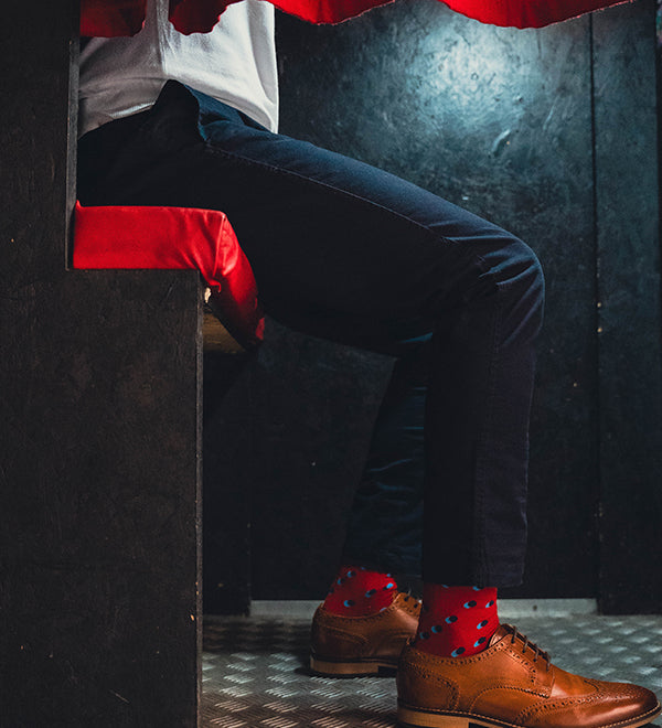 Person wearing brown shoes with red spot socks and dark pants peeking from behind a red curtain.