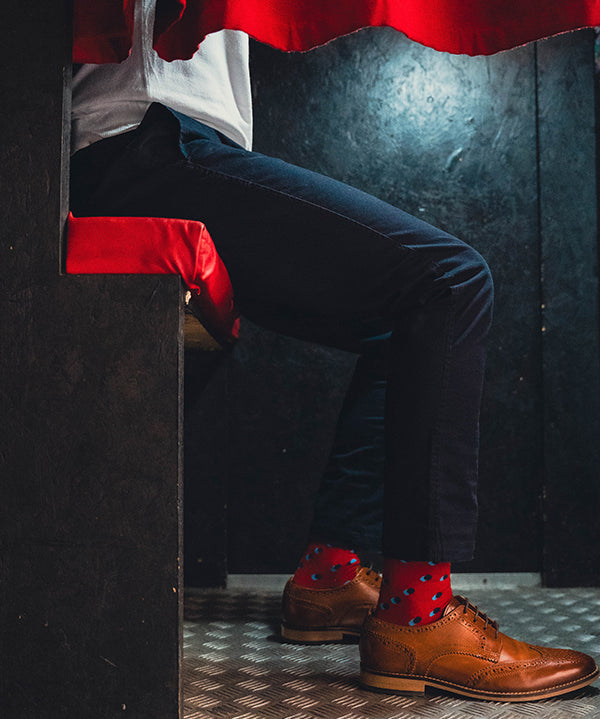 Person wearing brown shoes with red spot socks and dark pants peeking from behind a red curtain.