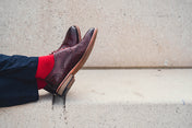 Brown dress shoes with red socks and a red shirt against a concrete wall.