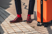 Person wearing brown shoes and red socks standing next to an orange suitcase on a paved walkway.