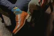Brown leather shoe with a colorful sock worn by a person sitting on a stone bench.