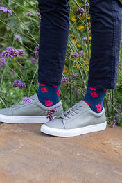 Person wearing blue socks with red floral patterns and gray sneakers standing on a rock with greenery in the background.