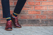 Person wearing burgundy shoes and colourful crew socks standing against a brick wall.