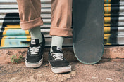 Person wearing black Vans shoes with white soles and mint green socks, standing next to a skateboard on a street.