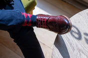Person wearing a dark shoe and red and black diamond design sock on a wooden floor.