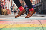 Person jumping on a rainbow crosswalk with brown shoes and red socks.