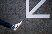 Person wearing white sneakers and bold stripey socks with a blue swoosh logo stepping onto a white arrow on a black asphalt surface.