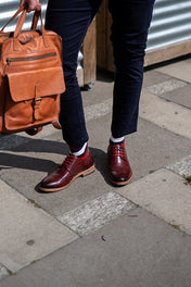 Person wearing brown leather shoes with striped socks and holding a brown leather bag on a sidewalk.